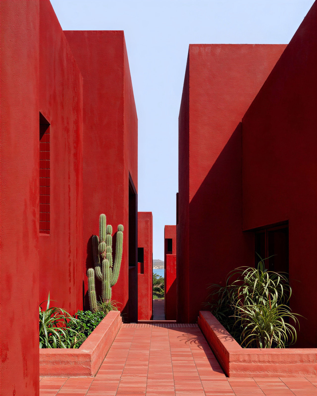 Passage between red volumes with cacti and plants, opening onto the sea