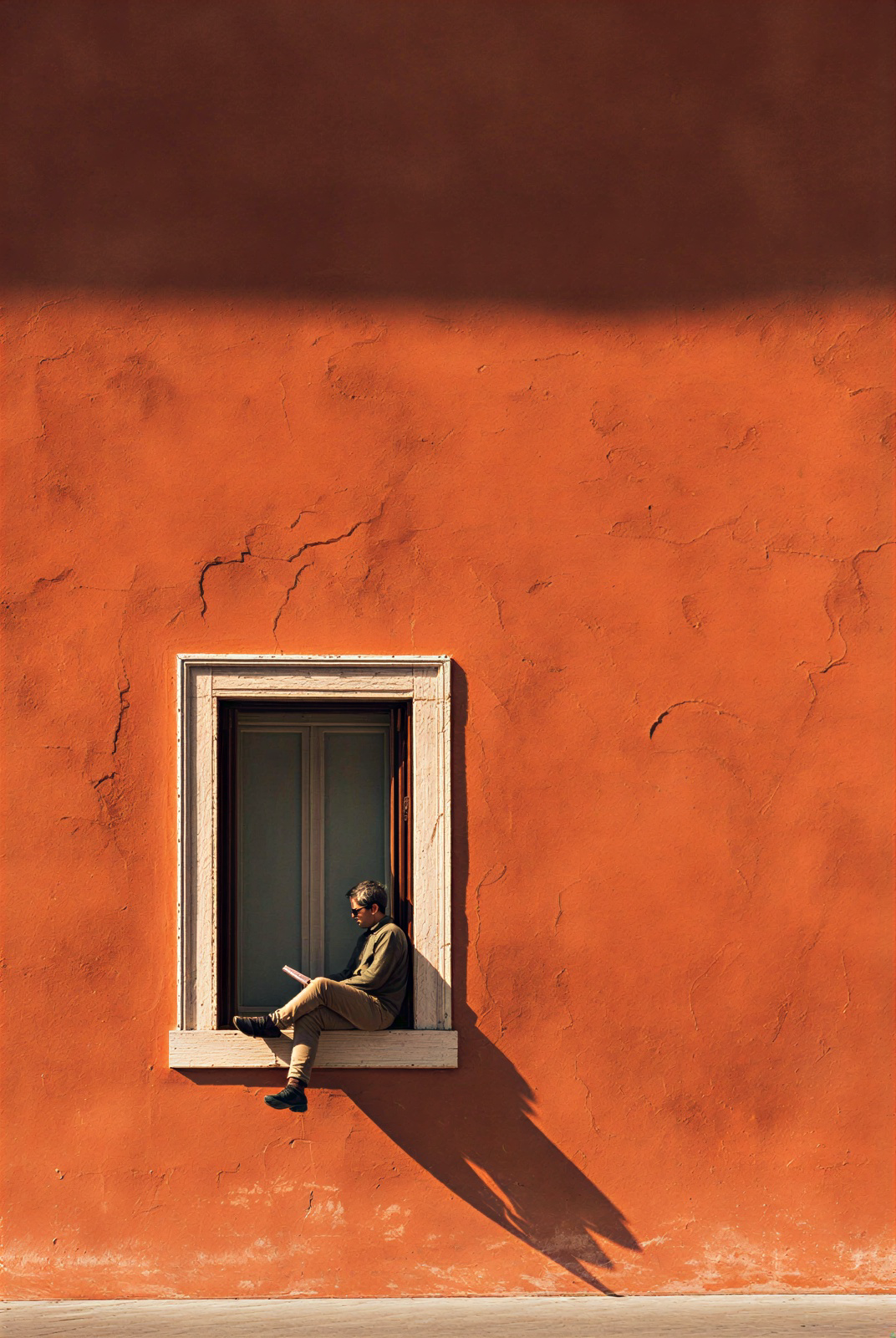 A person reading on the sill of a window in a terracotta wall, casting a long shadow